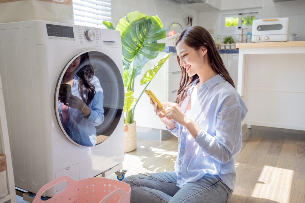 asian woman do the laundry in smart washing machine and use smartphone operate it at home