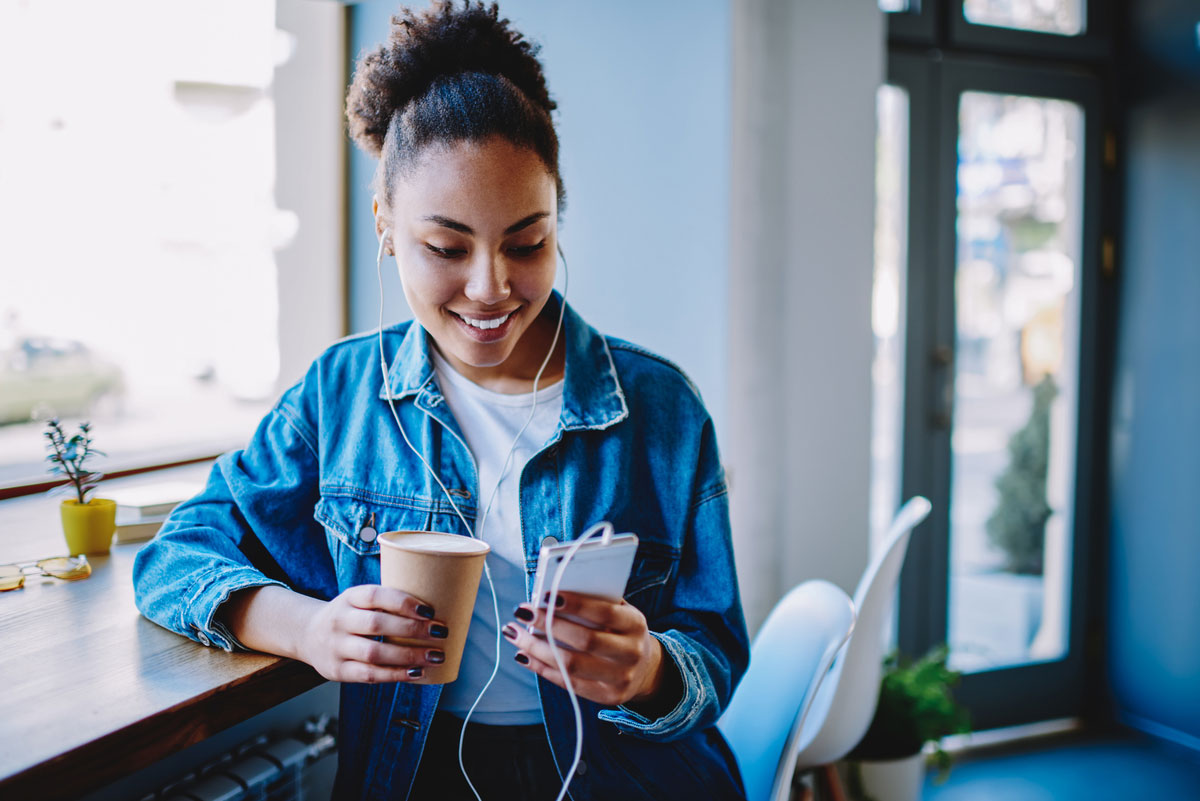 Young girl in a blue jean jacket wearing headphones holding a phone and a coffee at a cafe
