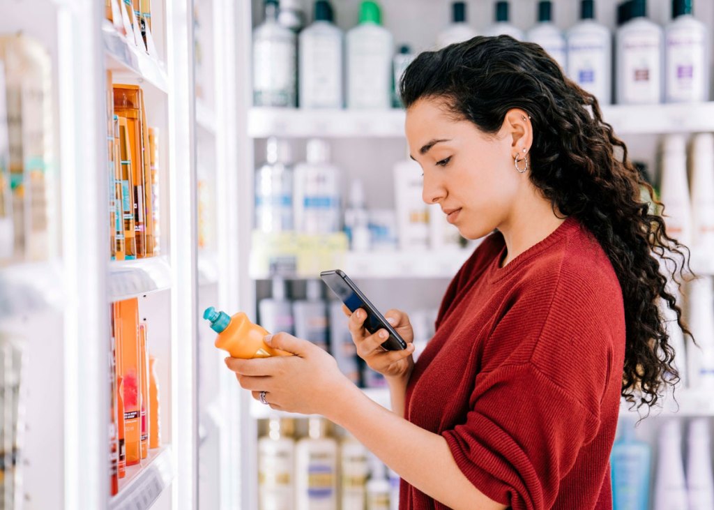 Woman with dark hair and red shirt holding phone looks at product in a pharmacy store